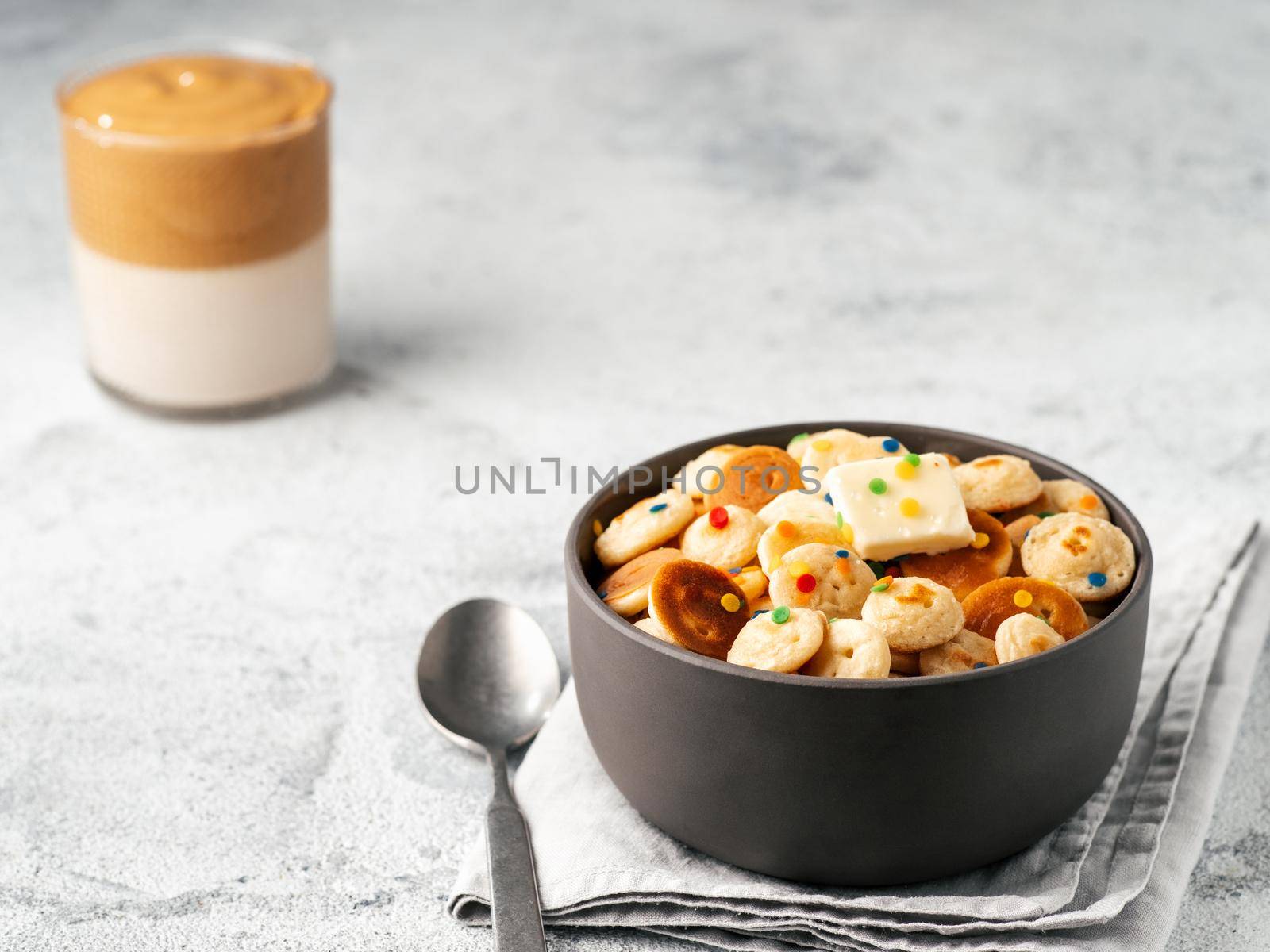 Mini pancakes cereal and dalgona coffee on gray cement background, copy space. Trendy food and drink - tiny panckakes served sprinkles and whipped instant coffee with milk or korean dalgona coffee