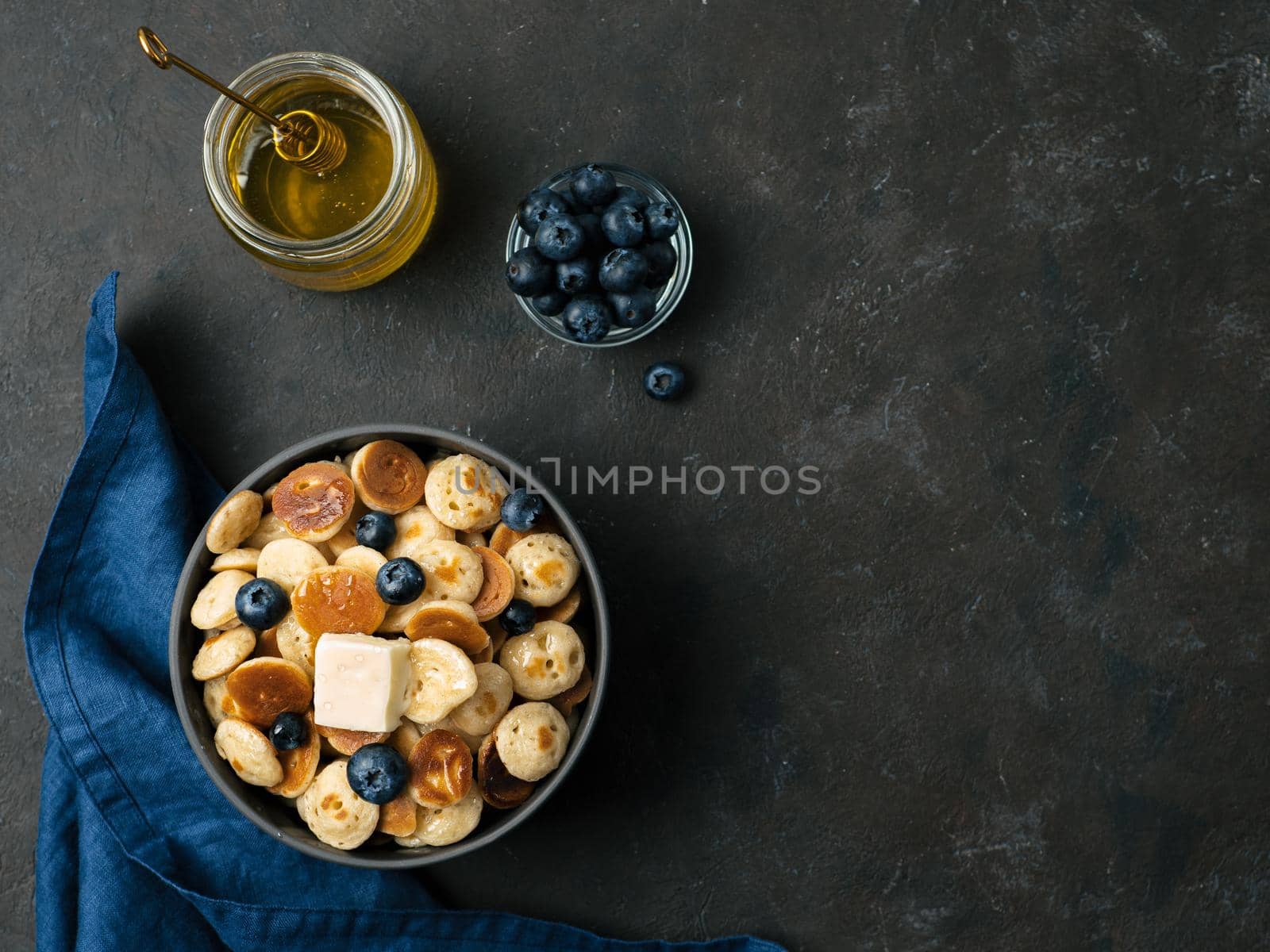 Trendy food - pancakes cereal served butter and blueberries. Heap of mini cereal pancakes in boul on dark background. Copy space right for text. Top view or flat lay.