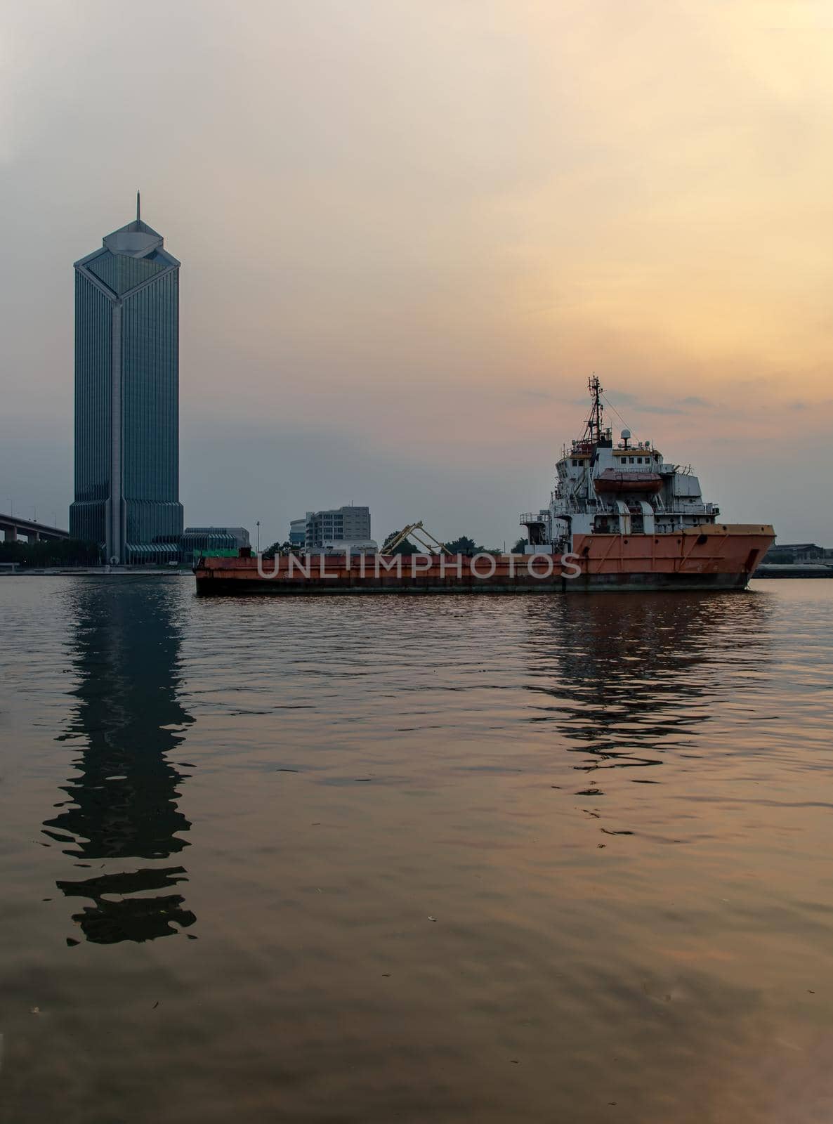 A cargo ship parked in the middle of the Chao Phraya River at evening with Modern building at the background. Selective focus.