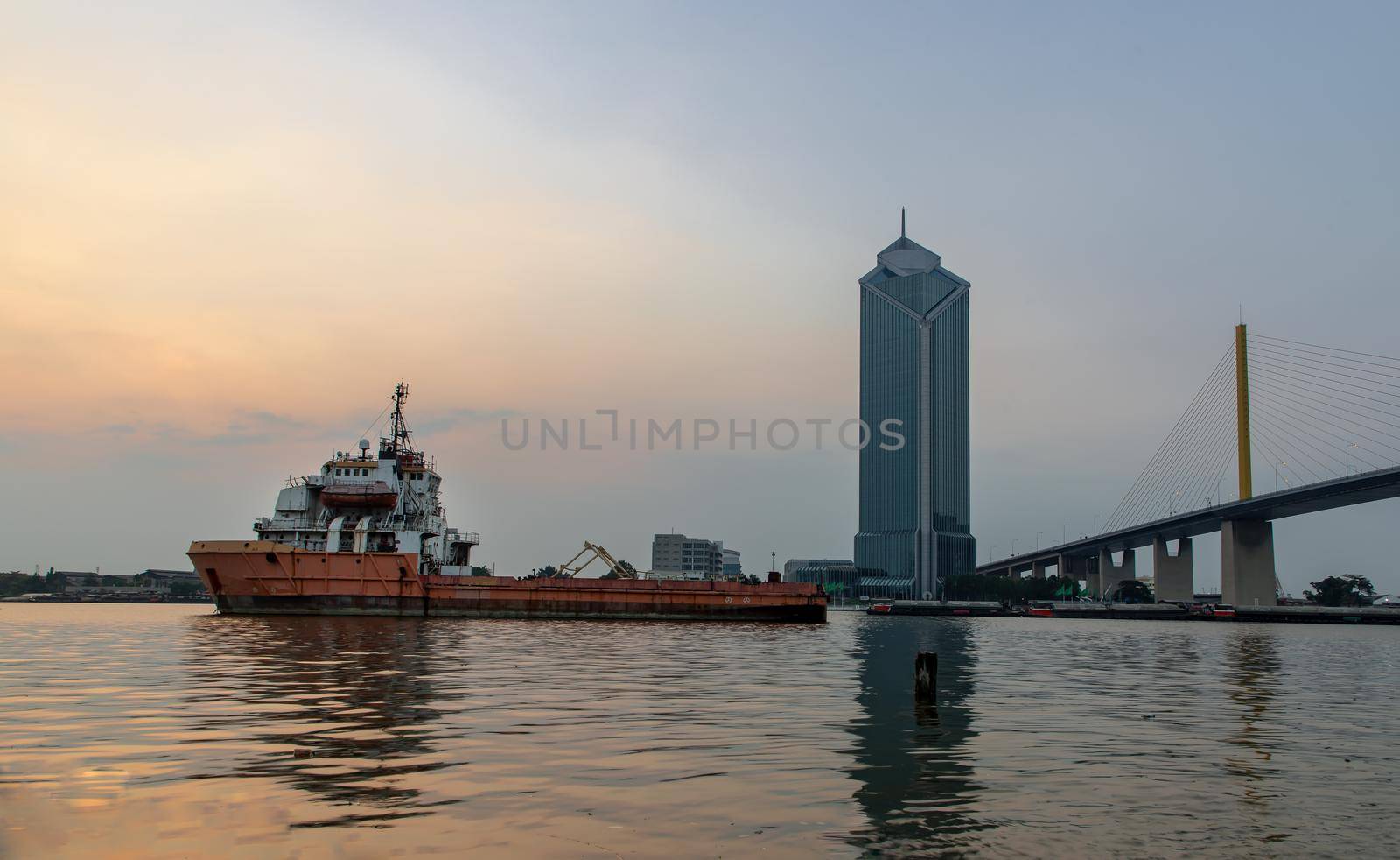 A cargo ship parked in the middle of the Chao Phraya River at evening with Modern building at the background. Selective focus.