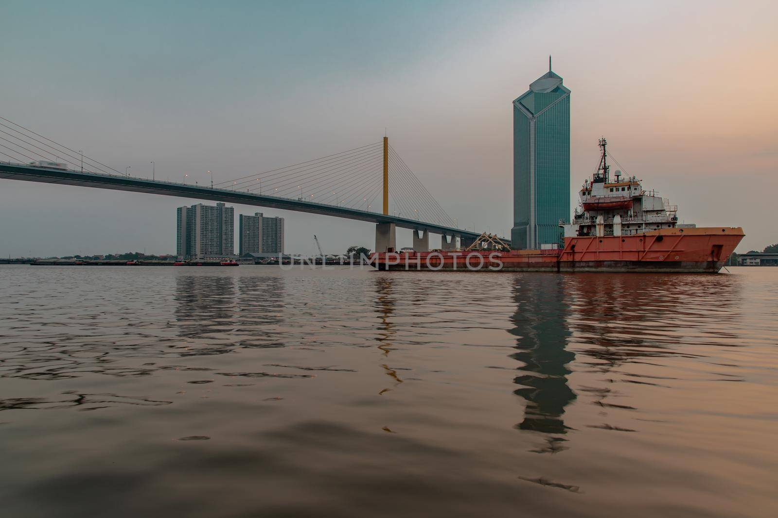 A cargo ship parked in the middle of the Chao Phraya River at evening with Modern building at the background. Selective focus.