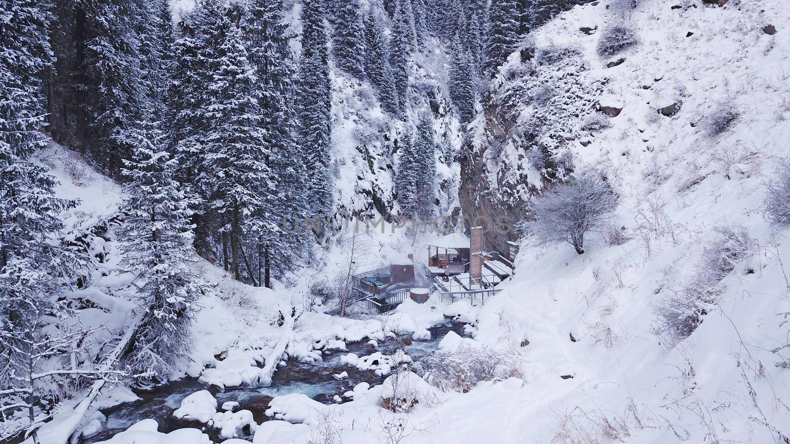 Hot springs in the snowy forest and mountains. Top view from the drone on the white gorge with the river. There are barrels of radon water. There is steam, people are bathing. Almarasan, Almaty.