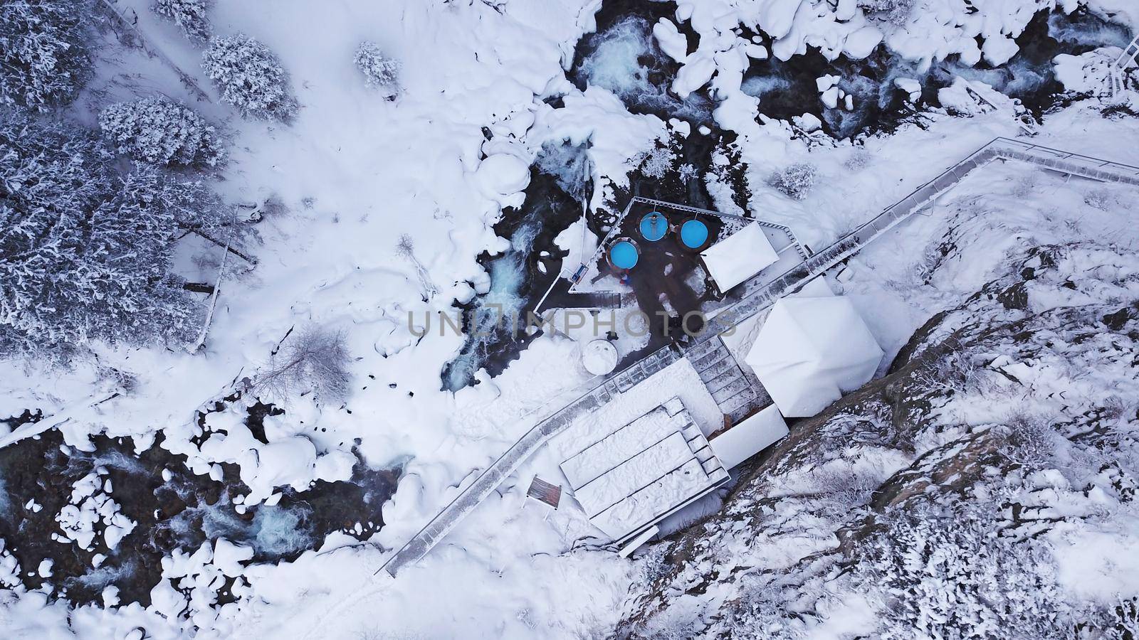 Hot springs in the snowy forest and mountains. Top view from the drone on the white gorge with the river. There are barrels of radon water. There is steam, people are bathing. Almarasan, Almaty.