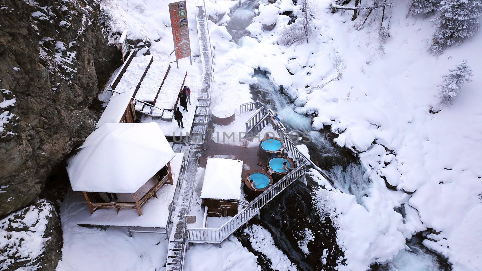 Hot springs in the snowy forest and mountains. Top view from the drone on the white gorge with the river. There are barrels of radon water. There is steam, people are bathing. Almarasan, Almaty.