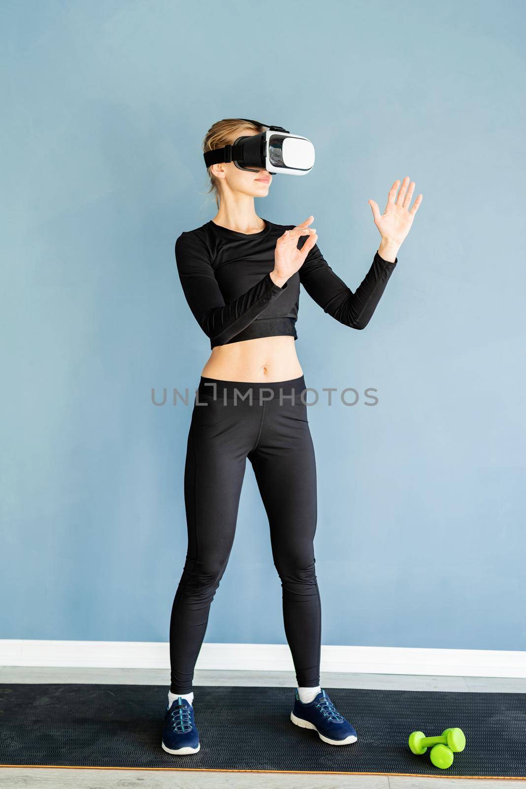 Young blonde woman in sport clothes wearing virtual reality glasses standing at fitness mat using VR interactive menu by Desperada