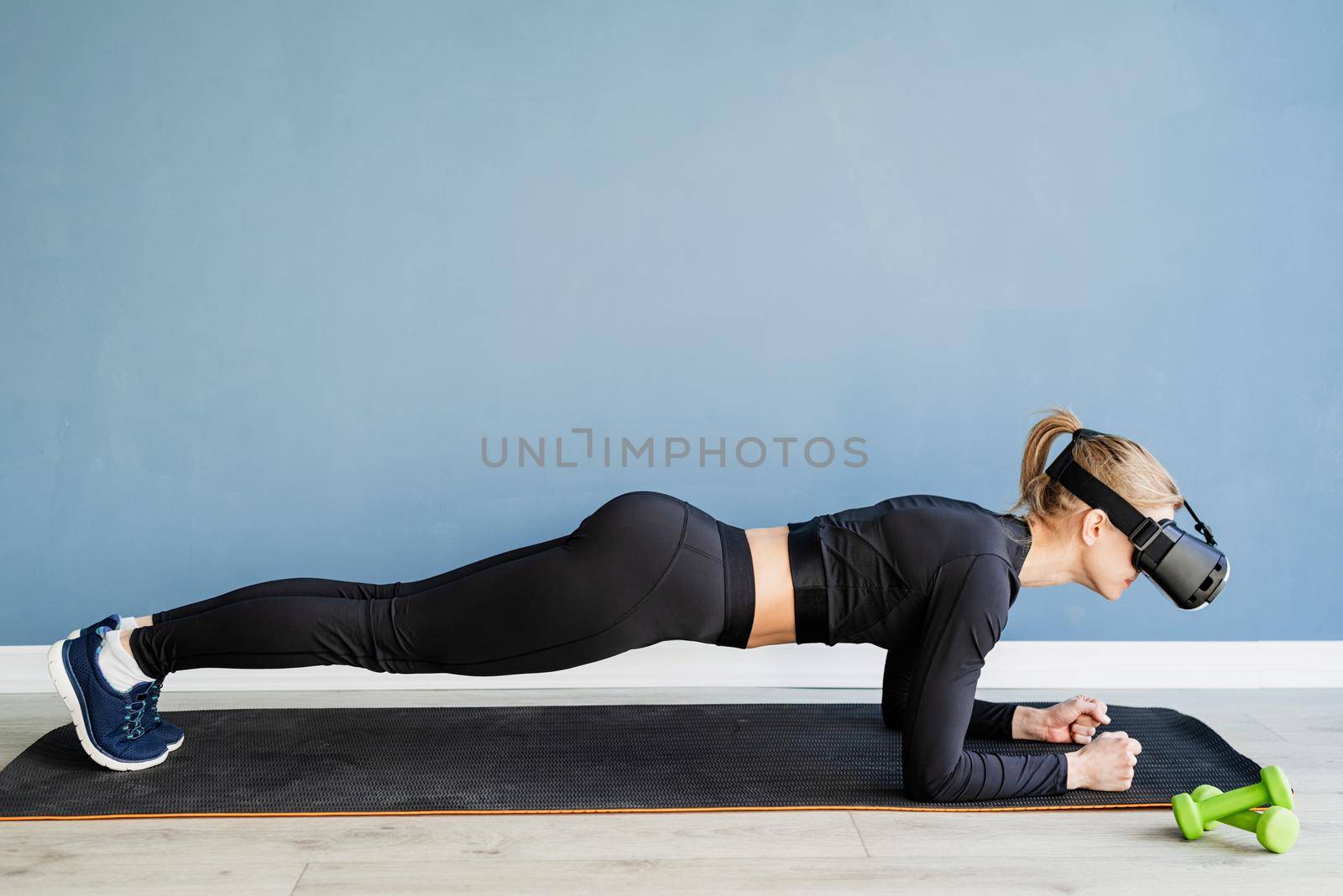 Fitness, sport and technology. Young athletic woman wearing virtual reality glasses doingplank at blue background