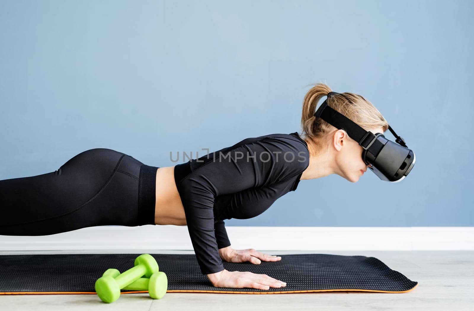 Young blonde woman wearing virtual reality glasses doing push ups at home by Desperada
