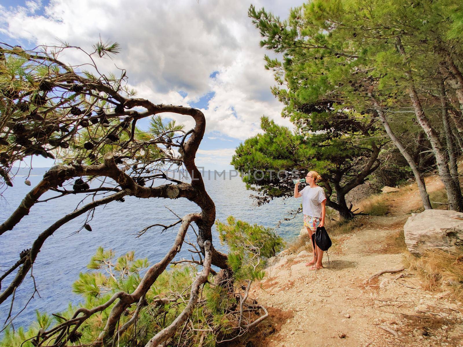 Young active feamle tourist taking a break, drinking water, wearing small backpack while walking on coastal path among pine trees looking for remote cove to swim alone in peace on seaside in Croatia by kasto