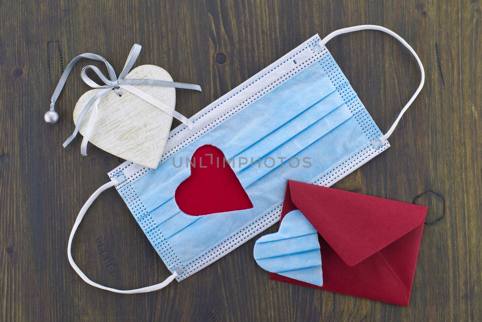 Face mask with cut out heart shape and red envelope on wooden background with free copy space. Social distancing and contact-less greetings