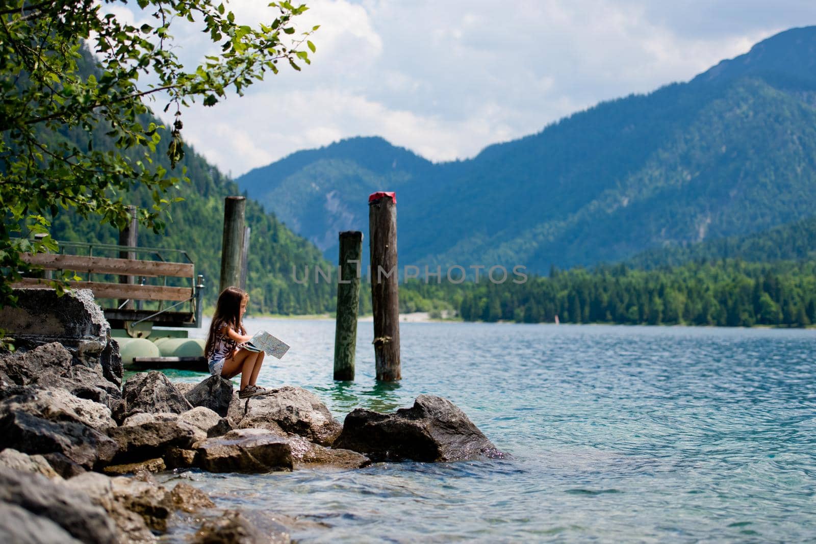 A little sweet girl sits on the shore of the Alpine lake Plansee and looks at the map. Plansee lake, Austria. by leonik