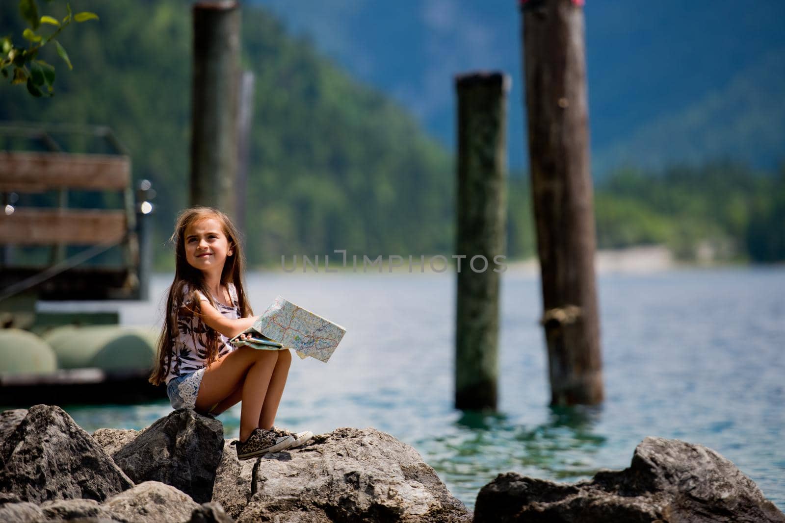 A little sweet girl sits on the shore of the Alpine lake Plansee and looks at the map. Plansee lake, Austria.