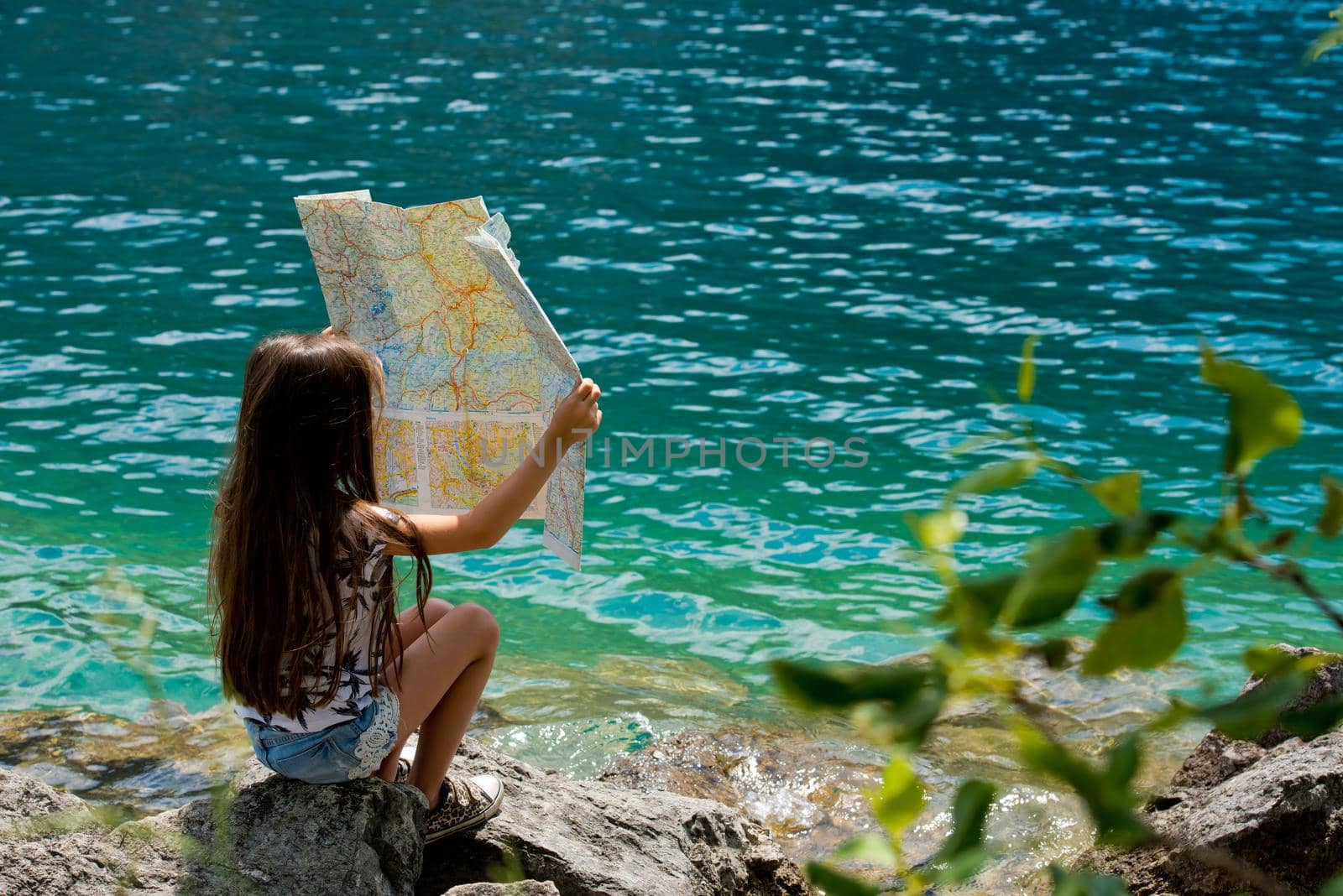 A little sweet girl sits on the shore of the Alpine lake Plansee and looks at the map. Plansee lake, Austria. Back view.