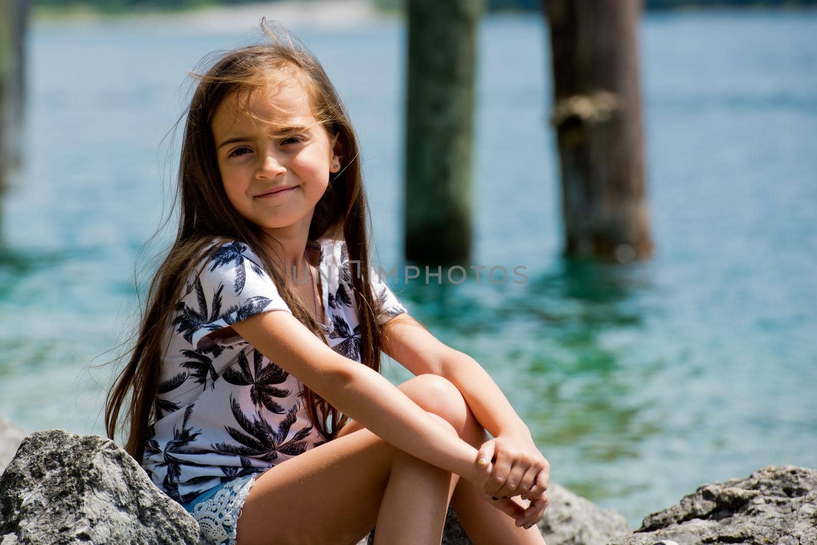 A little sweet girl sits on the shore of the Alpine lake. Plansee lake, Austria. Close-up.