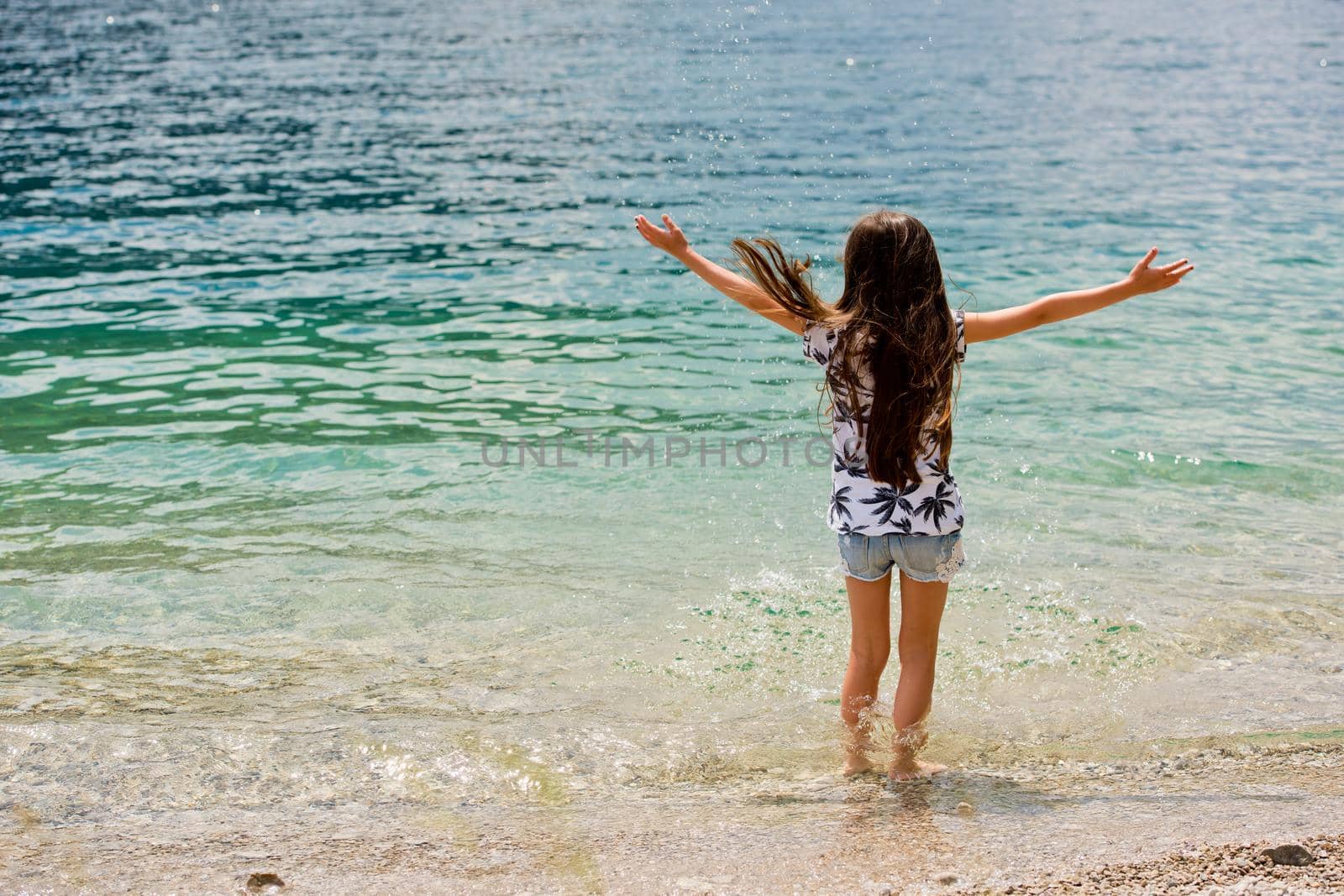 A cute little brunette girl stands on the shore of a lake Plansee with her arms outstretched in the distance. View from the back. by leonik