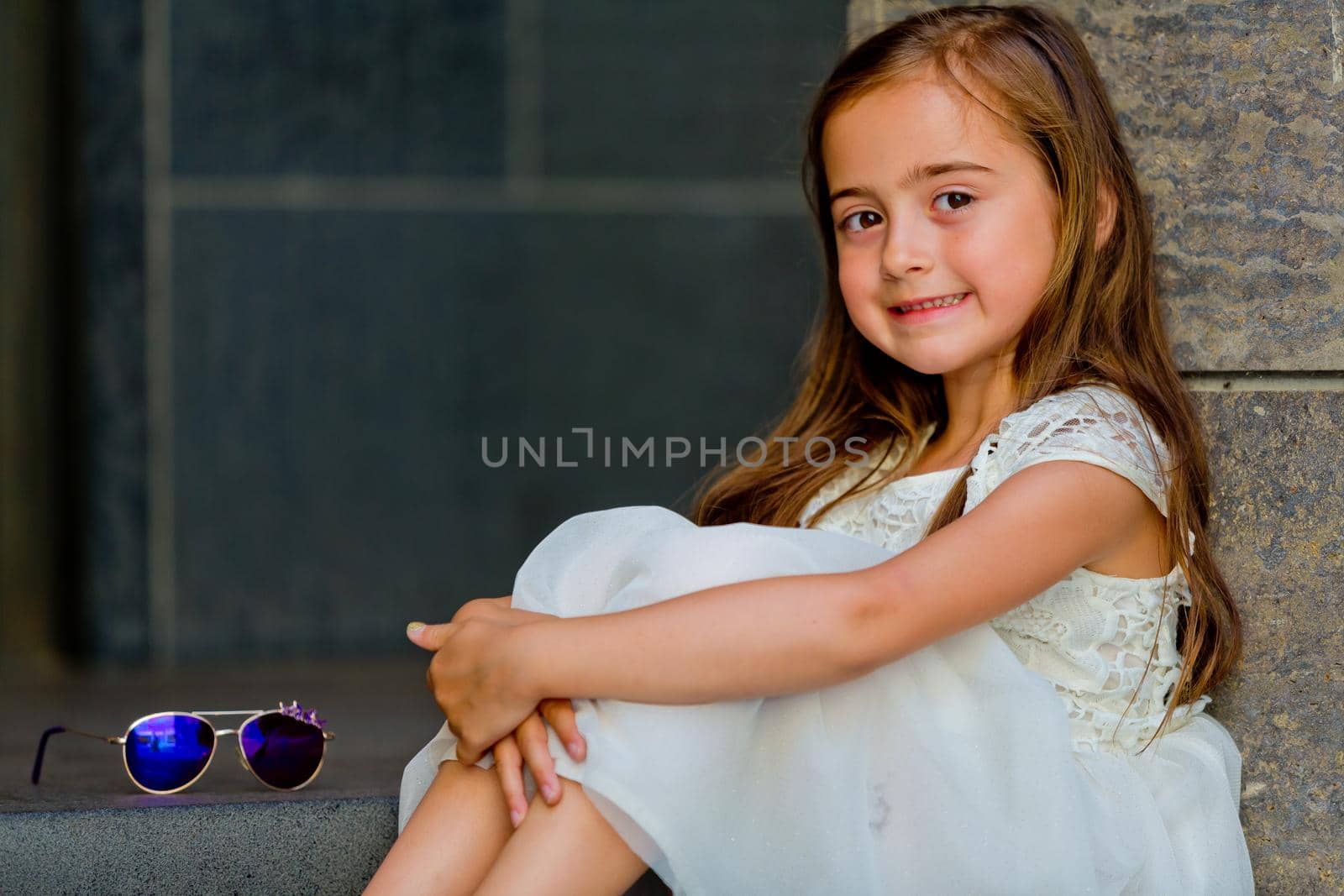 Little girl in a white dress is sitting on the steps and looking at the camera. Close-up.