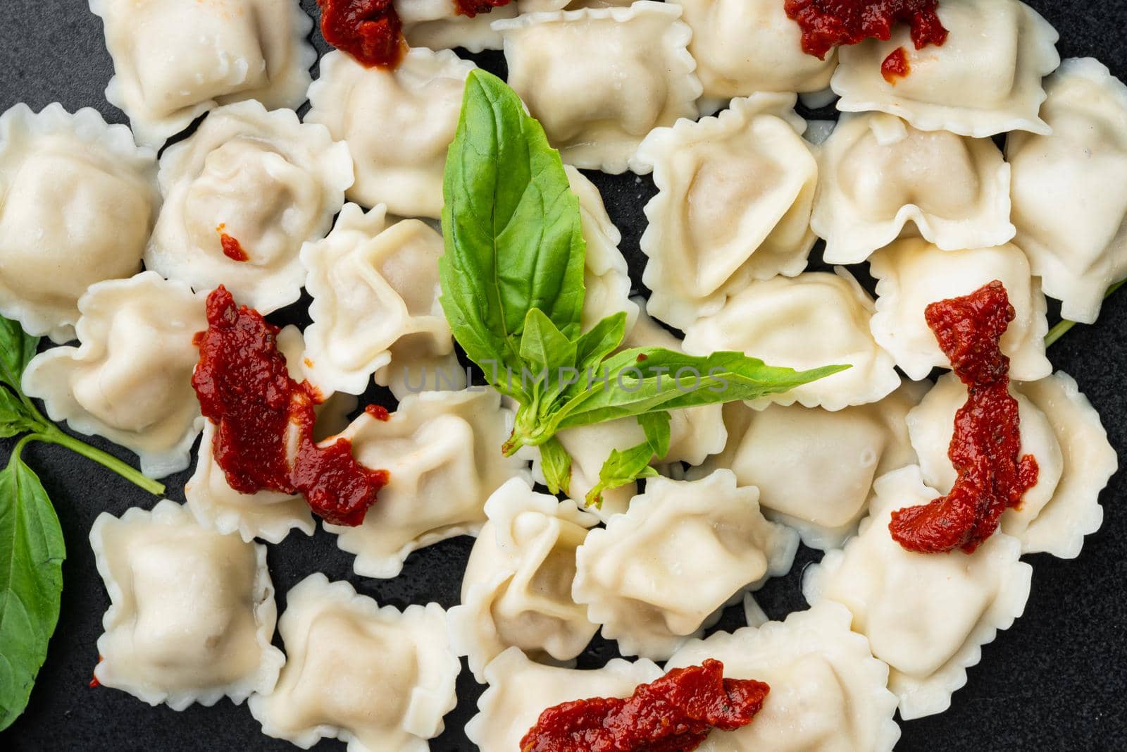 Appetizing ravioli with tomato sauce, cheese and basil set with basil parmesan and tomatoe on black plate, on black background