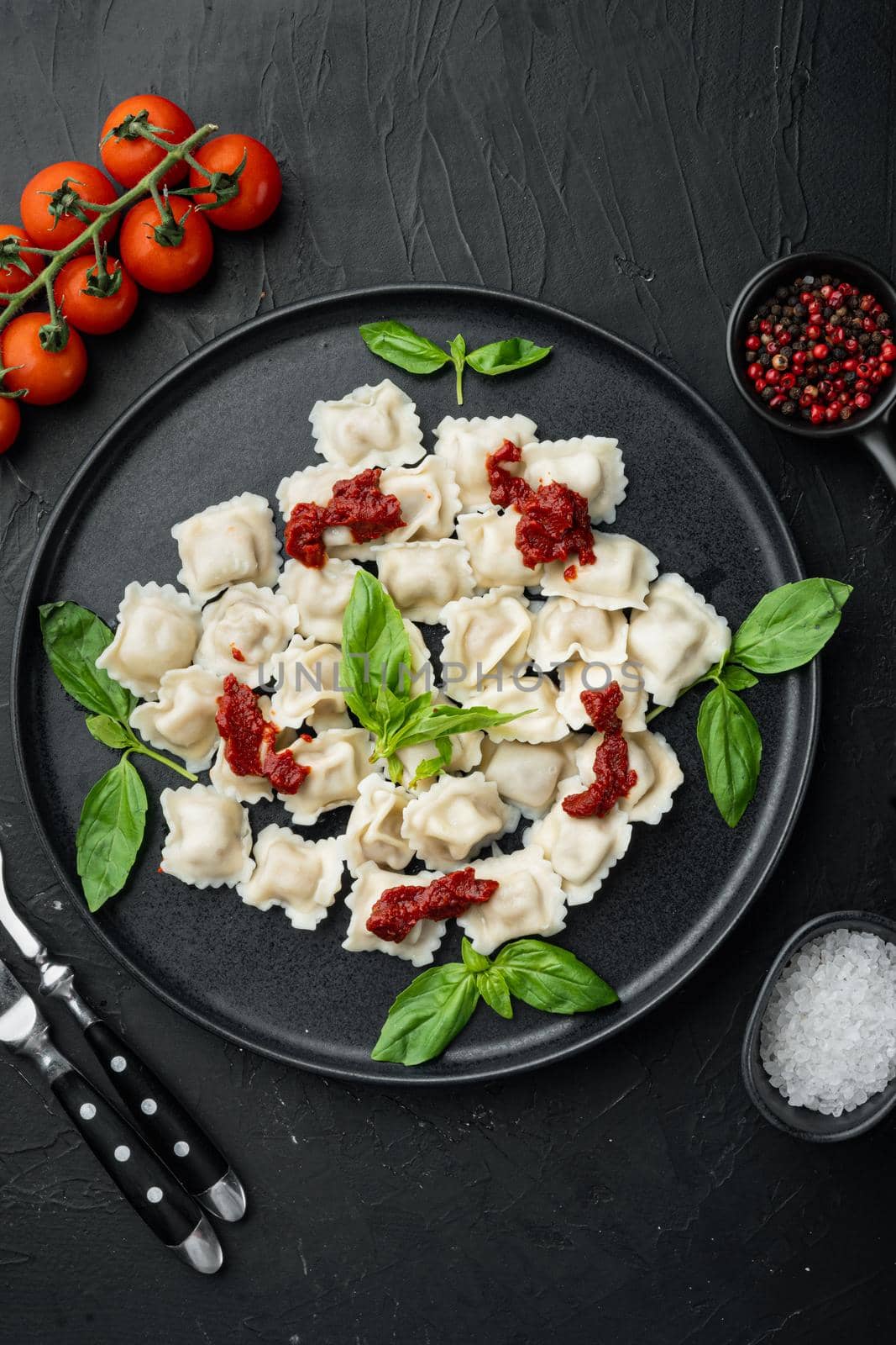 Appetizing ravioli with tomato sauce, cheese and basil set with basil parmesan and tomatoe on black plate, on black background, top view flat lay