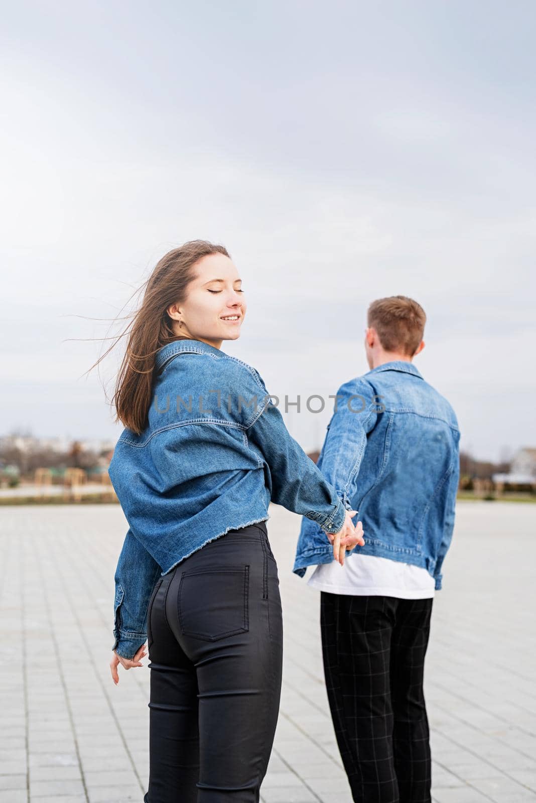 Young loving couple wearing jeans spending time together in the park having fun, woman holding hands with her boyfriend looking back