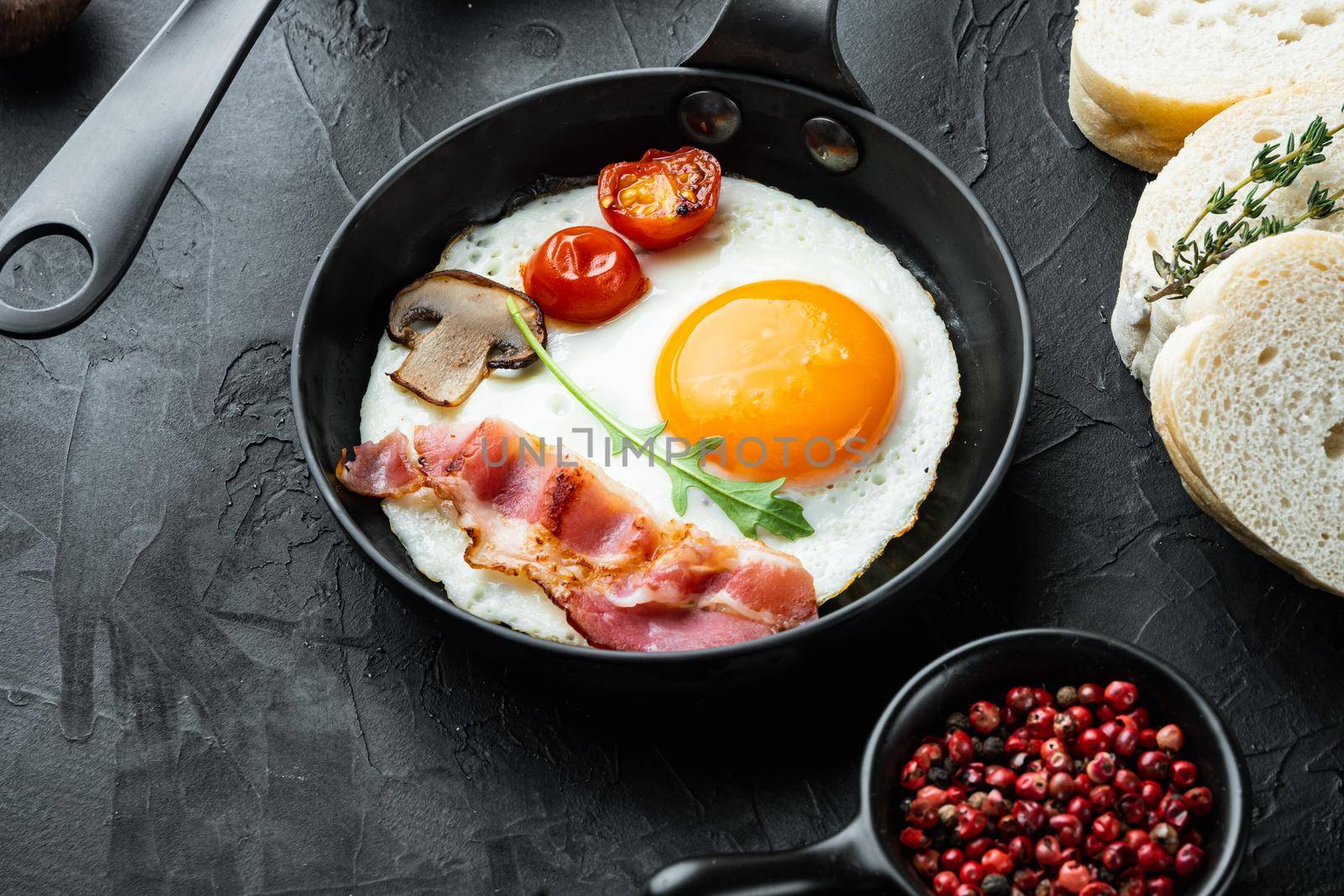 Fried eggs with bacon and vegetables set in cast iron frying pan, on black background