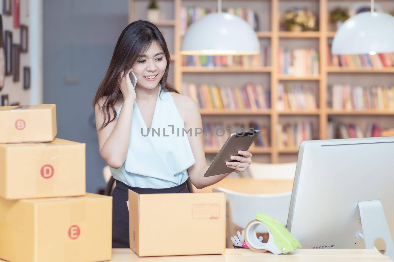 Young asian entrepreneur checking products stock on the smart tablet while talking to her customer at home office by Nuamfolio