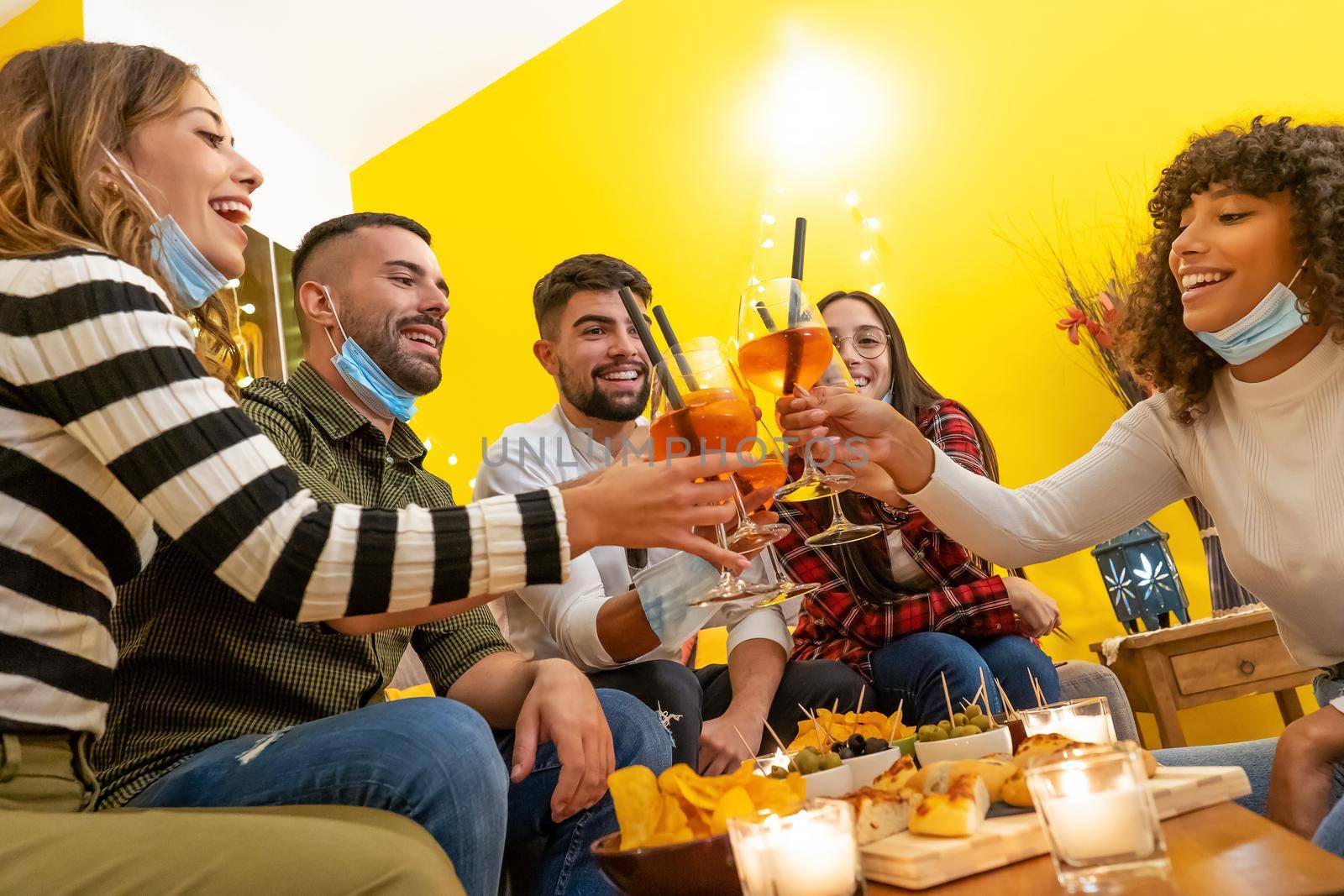 Group of young mixed race people celebrate at home wearing medical protection mask sitting at sofa around laden table. happy hour toasting at home with friends with new normal habits. Focus on glasses