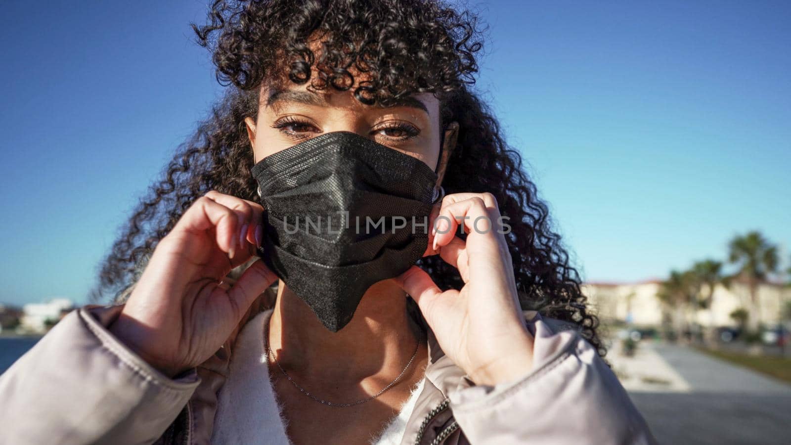 Close up portrait of young African American woman while wearing a black mask to avoid Coronavirus infection. Outdoor new normal social behaviors for human in the city against pandemic illness by robbyfontanesi