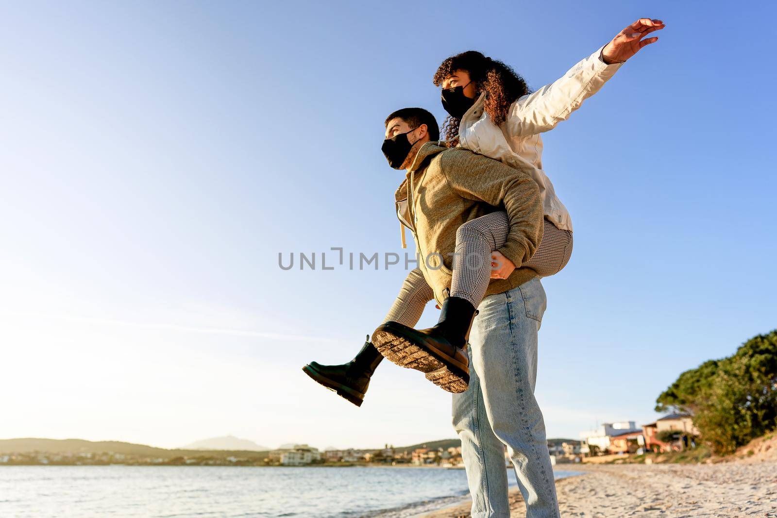 Happy young couple in love having fun doing girl on piggyback on the beach in winter vacations wearing Coronavirus protection mask at sunset. New normal human behaviors due the Covid pandemic risk