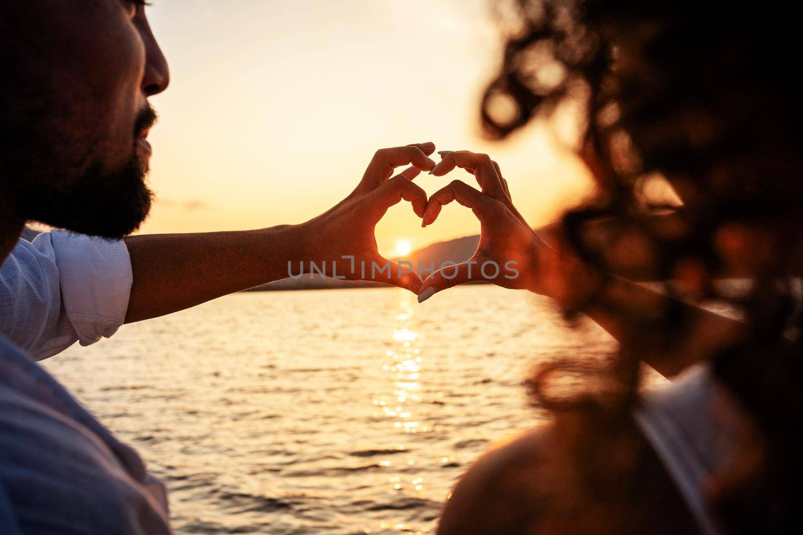 View from back of a young beautiful couple doing heart shape with fingers looking at the sun setting behind the mountains, reflecting light in the sea water. Valentine day love concept. Focus on hands by robbyfontanesi