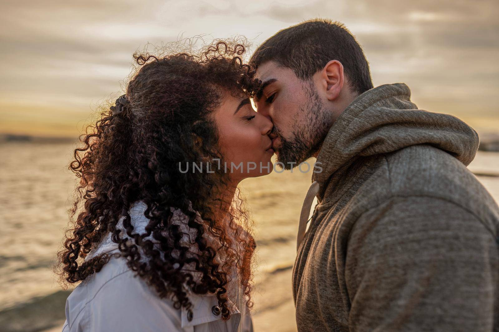 Head and shoulders portrait of young beautiful couple in love kissing at sunset in winter seaside resort with cloudy sky. Two millennials in vacation travel manifesting their heart sentiments outdoor by robbyfontanesi