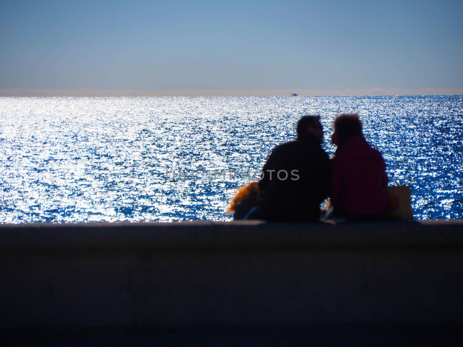 Blurred image of unrecognizable embraced lovers seen from behind sitting in front of the sea