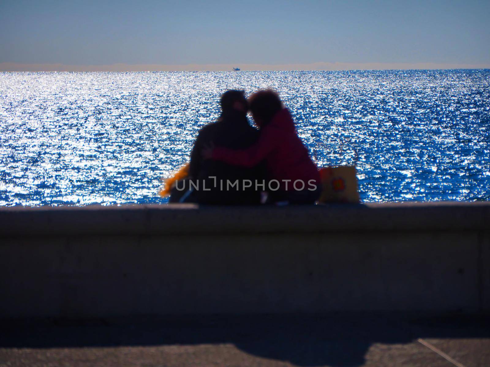 Blurred image of unrecognizable embraced lovers seen from behind sitting in front of the sea