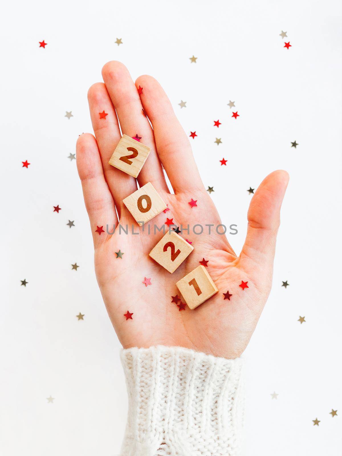 2021 on palm of hand. Woman in white knitted sweater holds wooden letters with stars confetti. Christmas and New Year background.