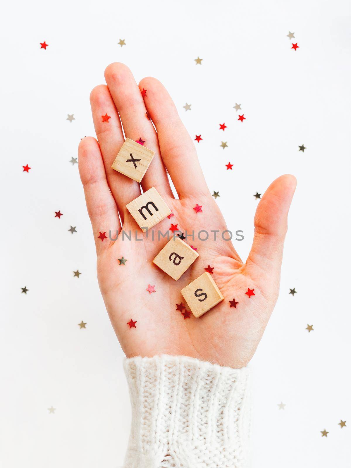 Word XMAS on palm of hand. Woman in white knitted sweater holds wooden letters with stars confetti. Christmas and New Year background.