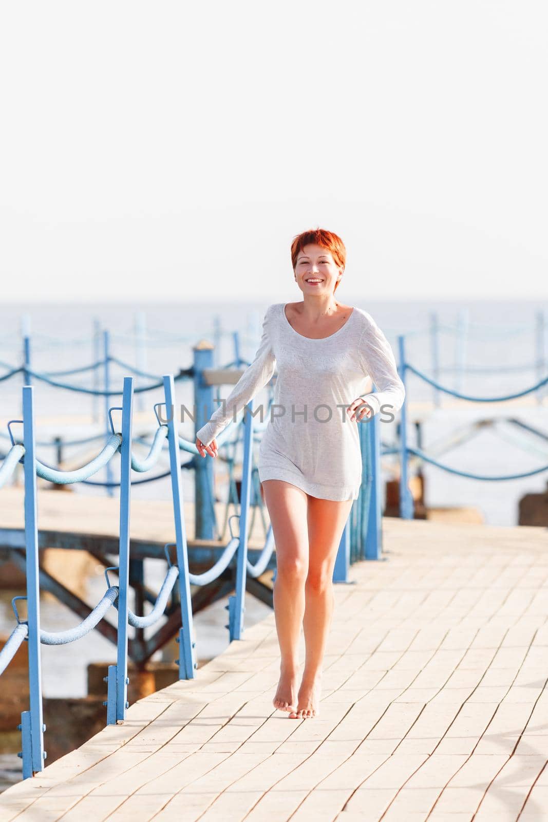 Wide smiling young woman with red short hair cut running on wooden pier. Coastal morning. Natural beauty. Happiness and power of youth.