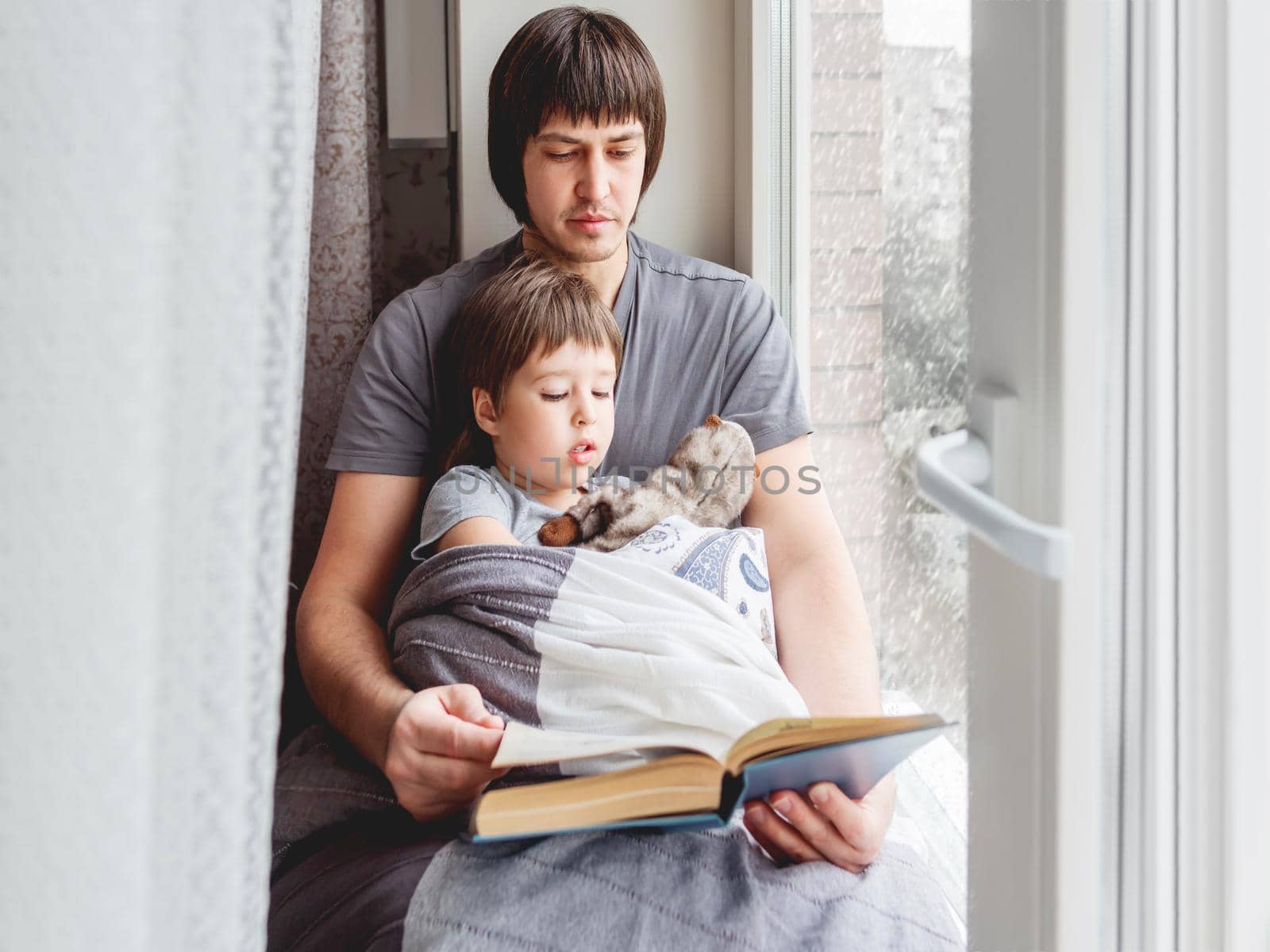Father reads book to his son. Cozy family time on windowsill while snowfall outside. Toddler boy sits together with fluffy toy cover himself with blanket. by aksenovko