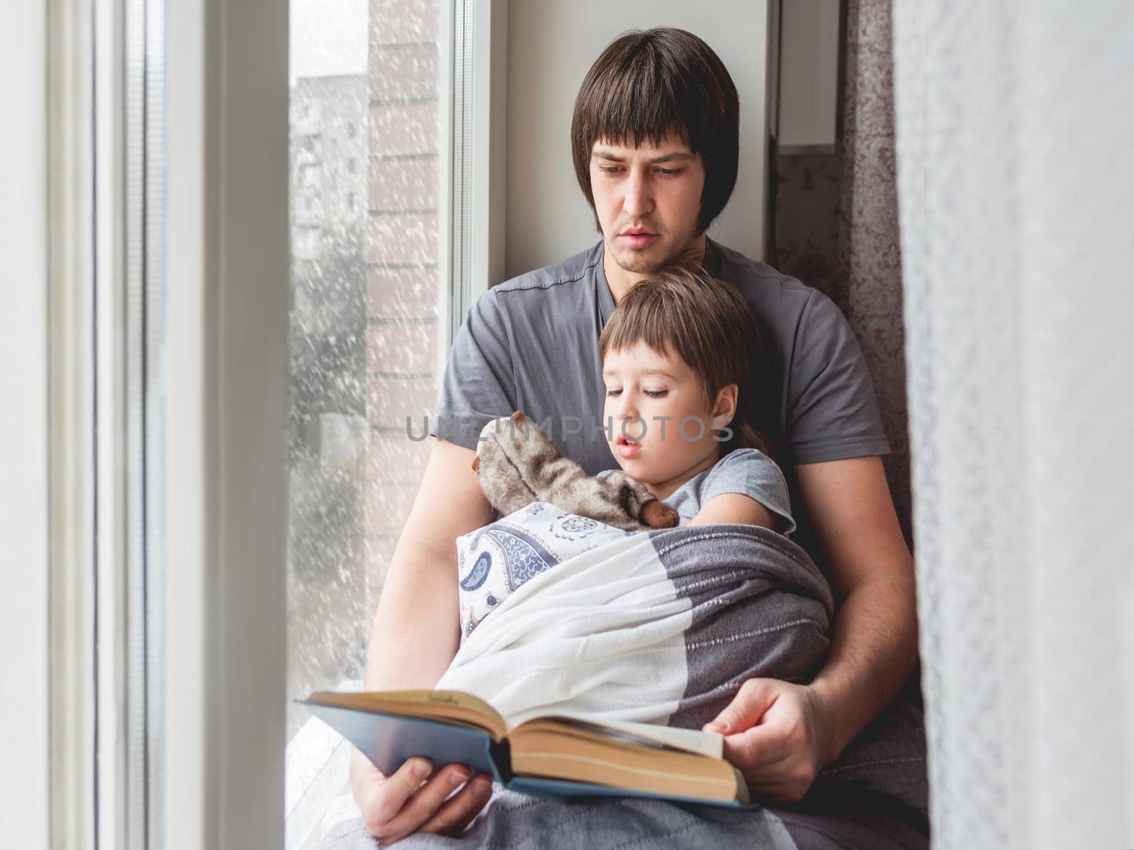 Father reads book to his son. Cozy family time on windowsill while snowfall outside. Toddler boy sits together with fluffy toy cover himself with blanket.