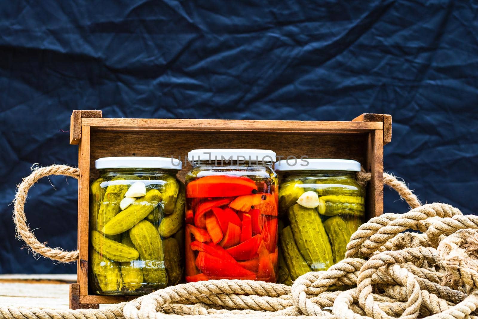 Wooden crate with glass jars with pickled red bell peppers and pickled cucumbers (pickles) isolated. Jars with variety of pickled vegetables. Preserved food concept in a rustic composition.