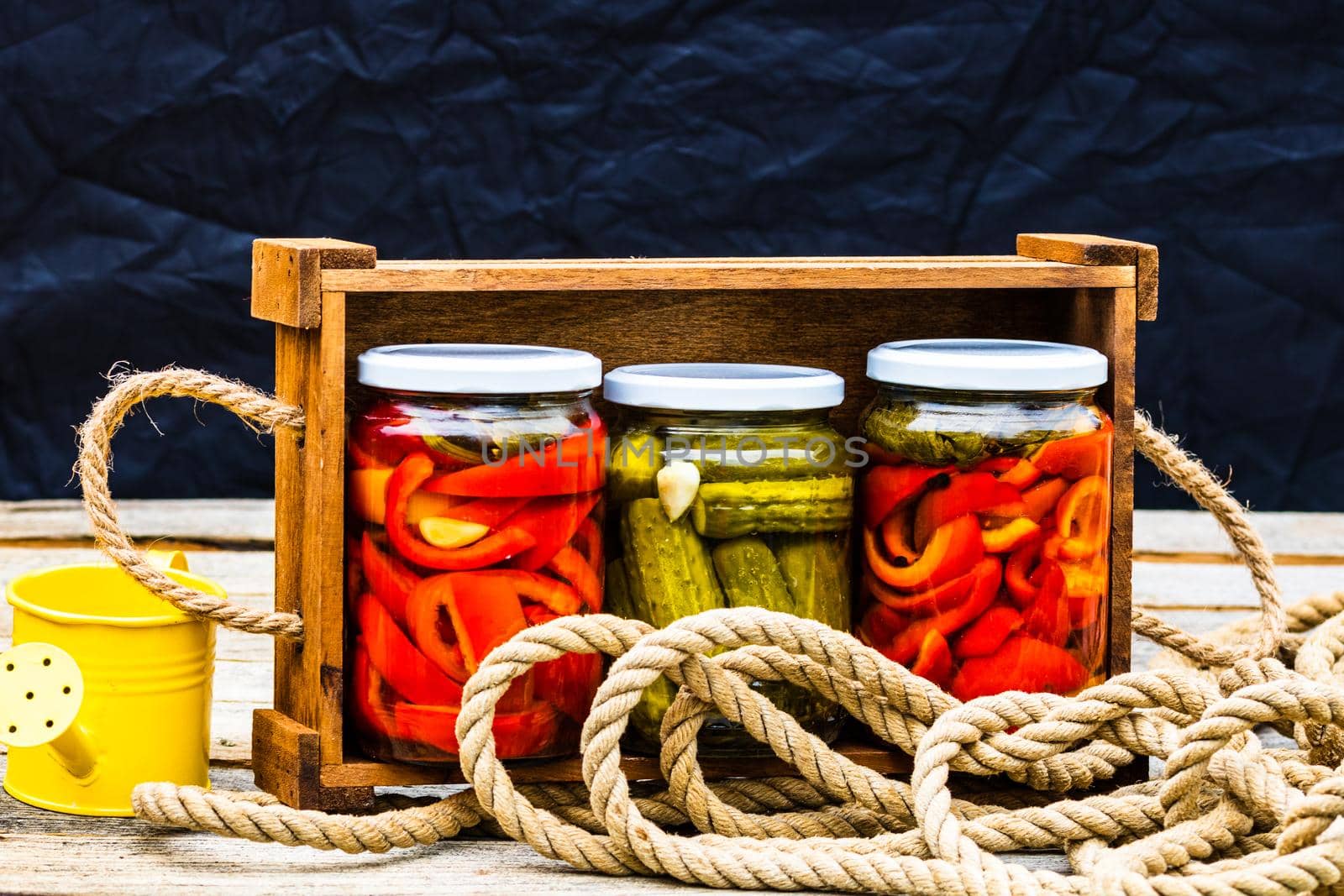 Wooden crate with glass jars with pickled red bell peppers and pickled cucumbers (pickles) isolated. Jars with variety of pickled vegetables. Preserved food concept in a rustic composition.