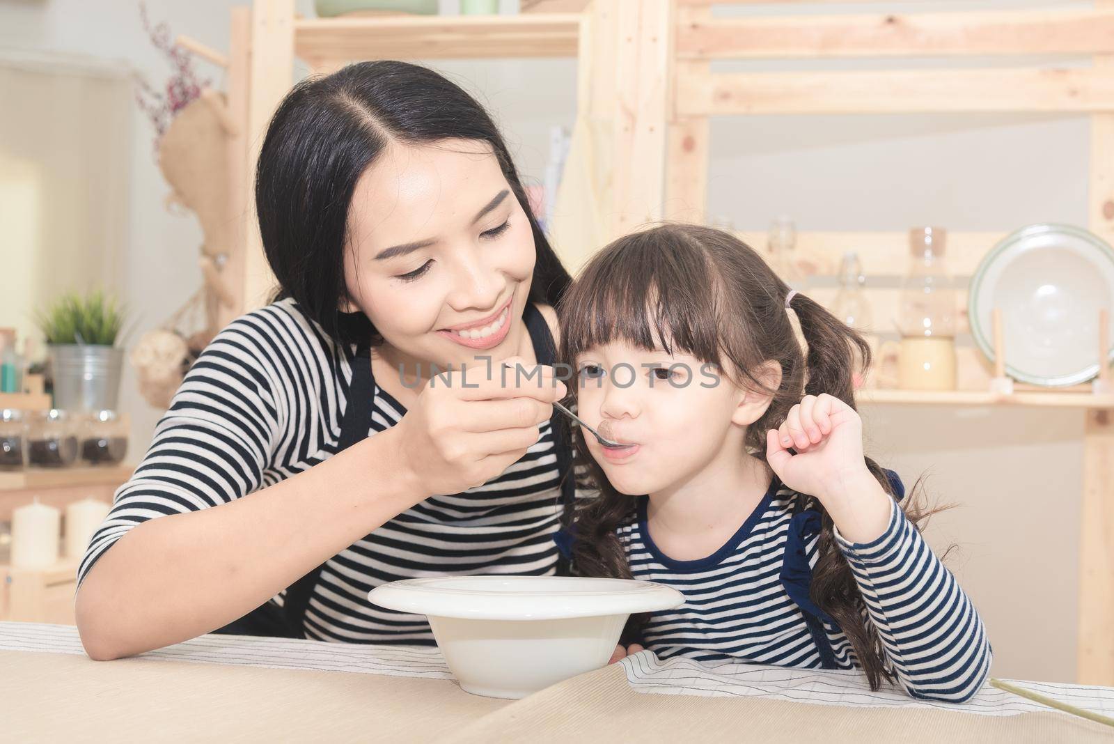 Happy family of asian mom feeding healthy breakfast to her cute daughter in the morning. by Nuamfolio