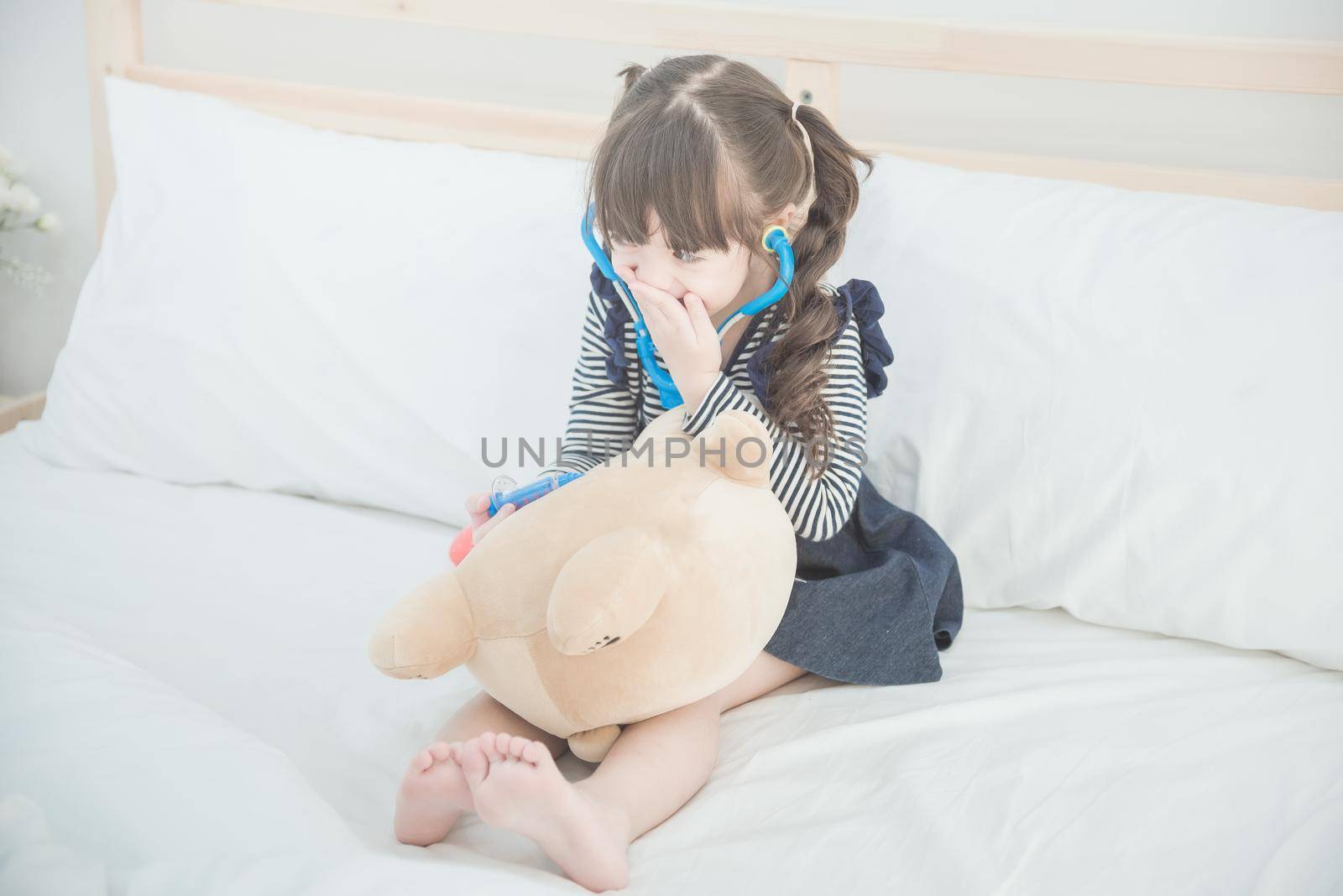 Cute asian little girl enjoy playing doctor with doctor toy set and cute doll while sitting on bed in kid's bedroom at home.