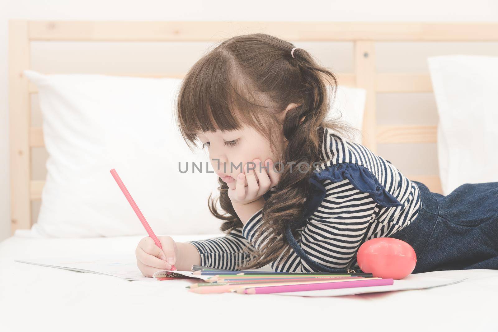 Cute asian little girl doing homework,writing with colourful pencils on bed at home. by Nuamfolio