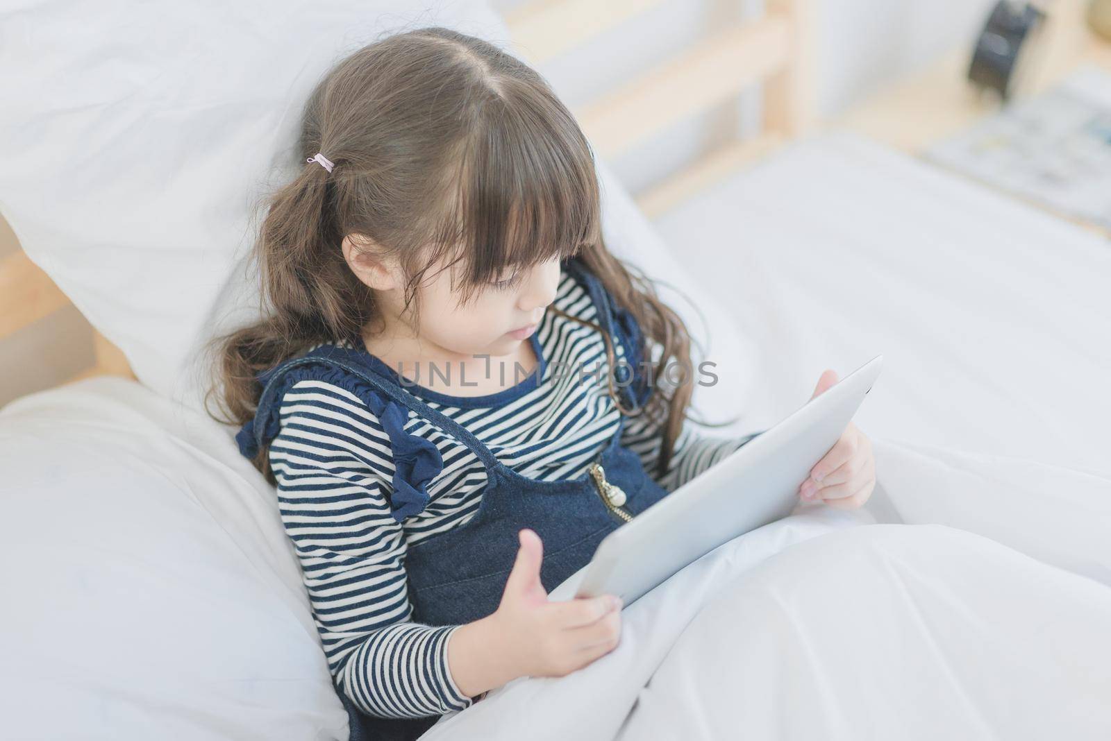 Cute asian little girl enjoy watching cartoon on smart tablet while sitting on bed in kid's bedroom at home.Photo for Happy Family concept.