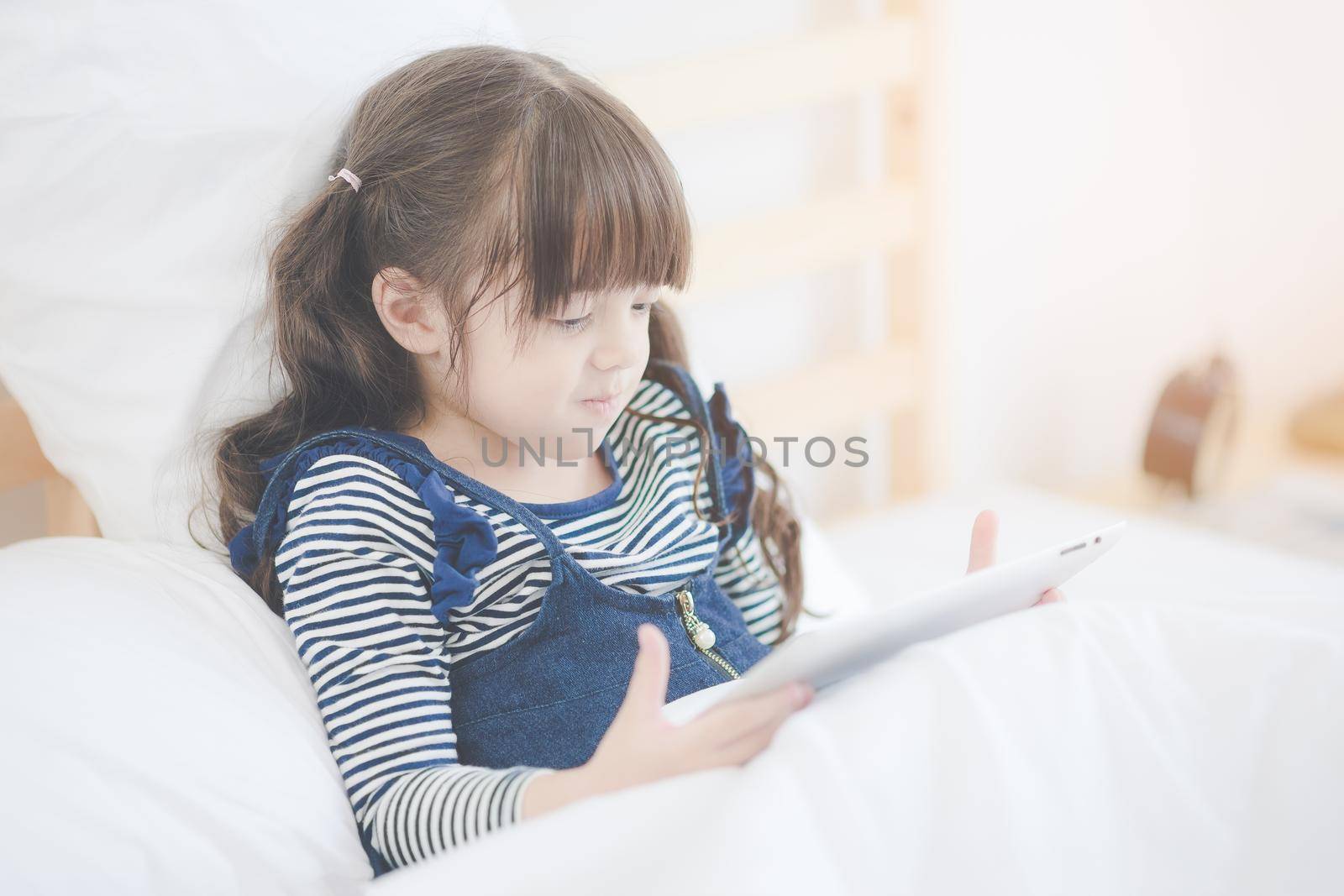 Cute asian little girl enjoy watching cartoon on smart tablet while sitting on bed in kid bedroom at home by Nuamfolio