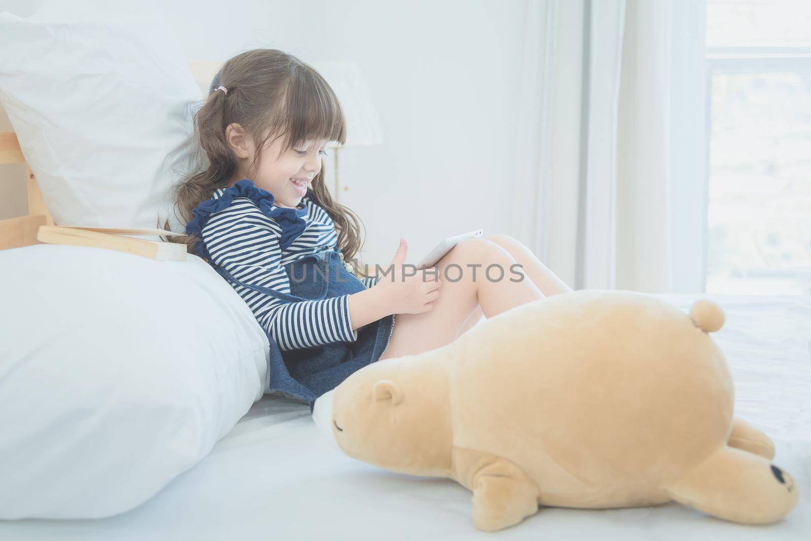 Cute little girl enjoy watching cartoon on smart tablet with cute doll while sitting on bed in kid bedroom at home