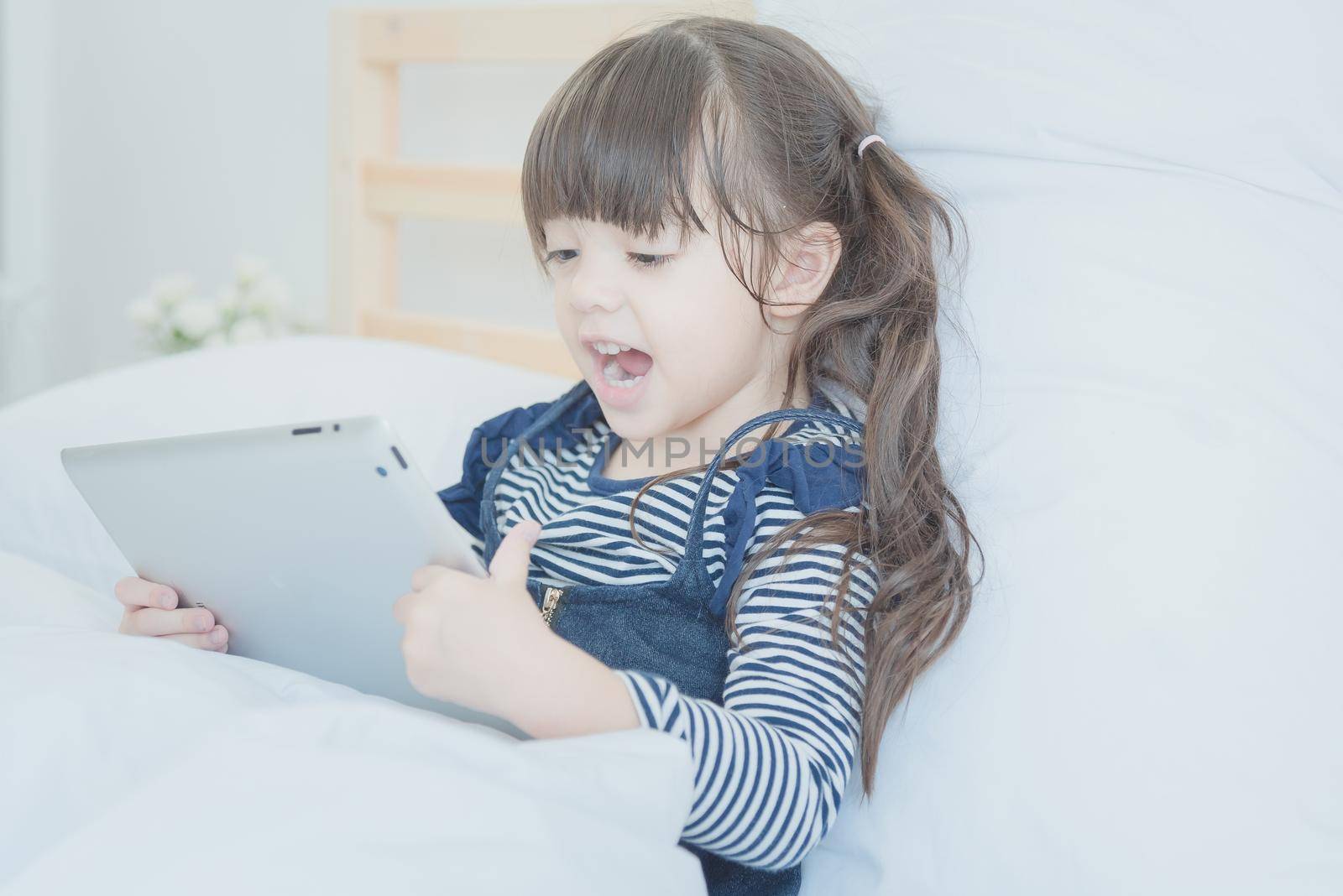 Cute little girl enjoy watching cartoon on smart tablet with cute doll while sitting on bed in bedroom at home by Nuamfolio