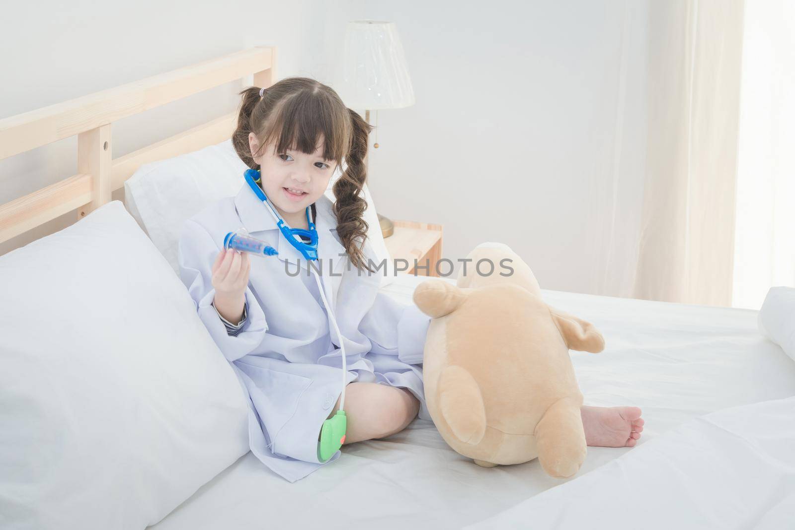 Cute little girl enjoy playing doctor with doctor toy set and cute doll while sitting on bed in bedroom at home by Nuamfolio