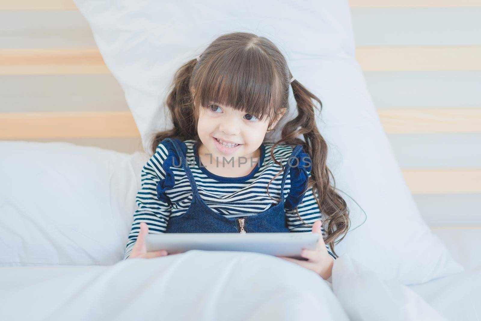 Cute kid girl smiling while using digital smart tablet on bed in kid's bedroom at home.Photo design for Happy Family and kids concept.