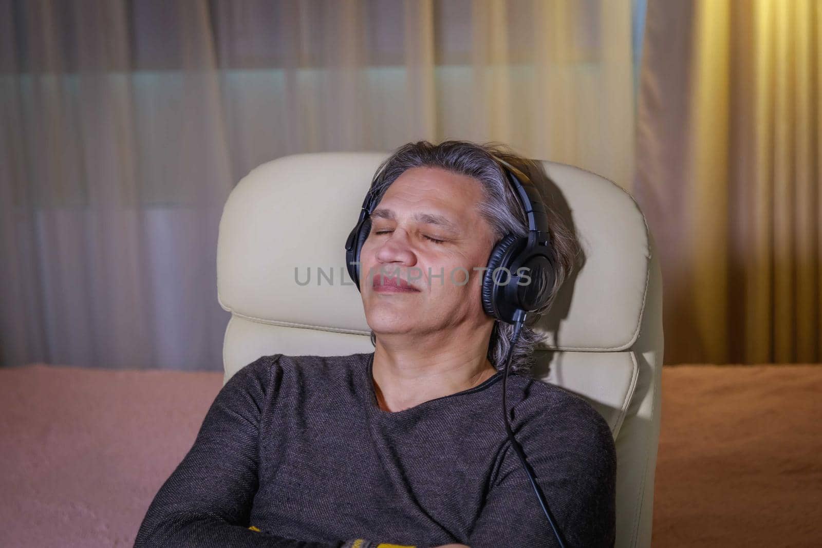 50-year-old man listens to music on headphones at home, sitting in a chair. by Yurich32