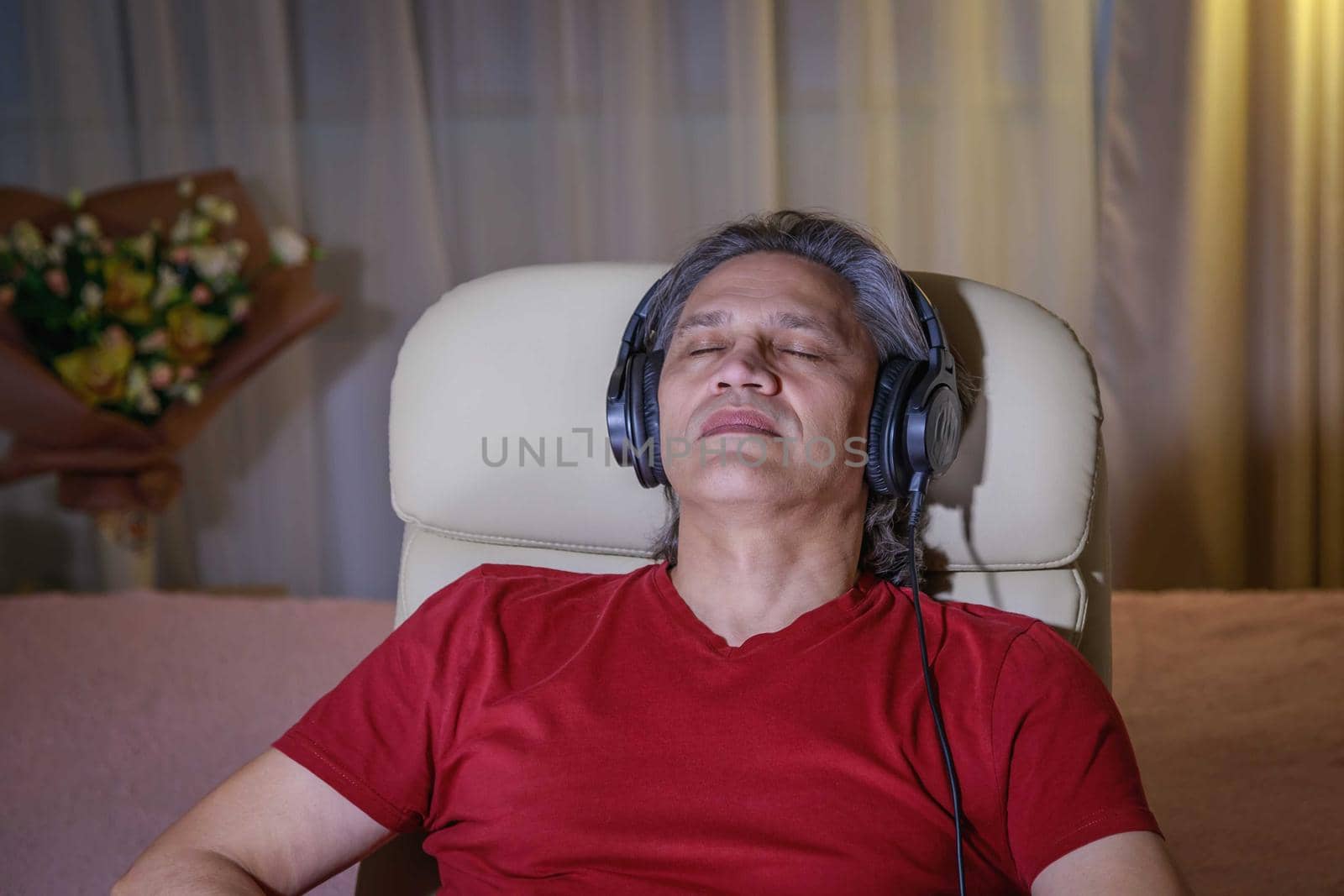 50-year-old man listens to music on headphones at home, sitting in a chair. Relaxing delight.