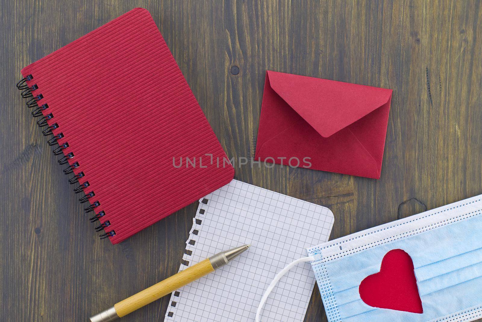 Medical mask with cut out heart shape alongside a wire bound notepad and pen and red envelope on wooden background with free copy space. Virus pandemic and quarantine concept